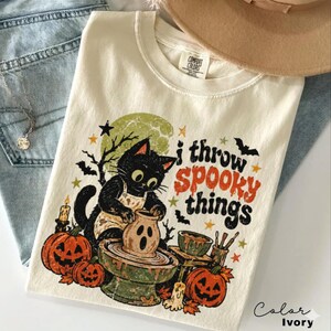 Op de afbeelding: Een ivoorkleurig t-shirt met een afbeelding van een zwarte kat in een schort die aan een pottenbakkersschijf een spookachtig kleifiguur vormt. Het ontwerp bevat oranje en zwarte tekst, pompoenen, vleermuizen en sterren.