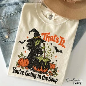 May include: An ivory t-shirt featuring a graphic of a green-skinned witch stirring a black cauldron. The text reads "That's It. You're Going in the Soup". The design includes orange pumpkins, black bats, stars, and a small potion bottle.