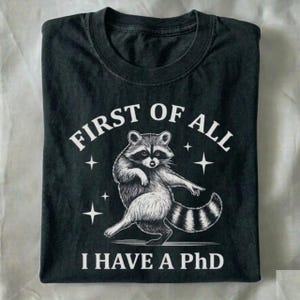 May include: Black t-shirt with a graphic of a raccoon in a dancing pose. The text "FIRST OF ALL" arches above the raccoon, and "I HAVE A PhD" is printed below. The design is in white.