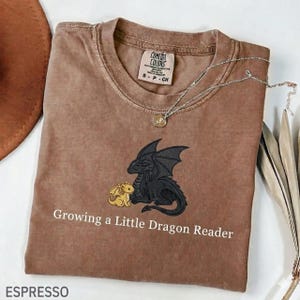 Może przedstawiać: Koszulka w kolorze espresso z motywem smoka. Projekt przedstawia czarnego smoka i mniejszego żółtego smoka. Tekst "Growing a Little Dragon Reader" znajduje się pod grafiką smoka. Srebrny naszyjnik z wisiorkiem leży na koszulce.