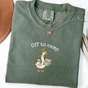 Op de afbeelding: Een olijfgroen T-shirt met ronde hals en een afbeelding van een gans met een vissershoedje, een rugzak, een waterfles en een kaart. De tekst "Off to camp" staat in het wit boven de gans gedrukt en er ligt een dunne gouden ketting op de kraag.