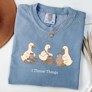 Könnte beinhalten: Ein blaues kurzärmeliges T-Shirt mit einer Grafik von drei weißen Enten, die an einer Töpferscheibe arbeiten, und dem in Weiß gedruckten Text I Throw Things. Das T-Shirt hat einen Rundhalsausschnitt und ein Comfort Colors Markenetikett am Kragen.