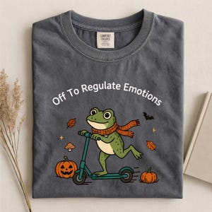 May include: A charcoal grey crew neck t-shirt featuring a graphic of a green frog wearing an orange scarf and riding a teal scooter. The text "Off To Regulate Emotions" is printed above the frog, surrounded by pumpkins, autumn leaves, and a bat.