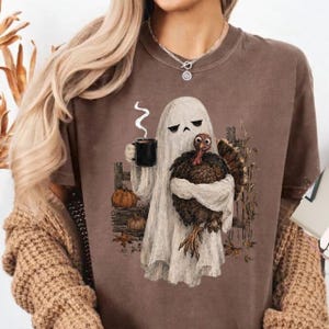 May include: A brown t-shirt featuring a graphic of a white ghost holding a black coffee mug and a brown turkey. The ghost stands in front of a wooden fence with two small pumpkins and autumn leaves at its base. The design has a muted colour palette.