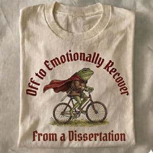 以下が含まれることがあります： クリーム色のTシャツ。自転車に乗ったカエルがマントと剣を身につけた、風変わりなイラストが描かれています。「Off to Emotionally Recover From a Dissertation」というテキストは、バーガンディ色です。
