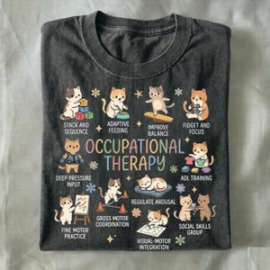 May include: Dark gray t-shirt featuring cartoon cats demonstrating occupational therapy activities. Text includes "Occupational Therapy" and phrases like "Stack and Sequence," "Adaptive Feeding," and "Fine Motor Practice."