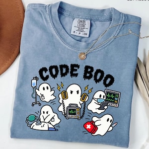 May include: A blue crew neck t-shirt featuring the text CODE BOO in black letters above a graphic of five white ghosts performing medical tasks, including using a defibrillator, an IV pole, a heart monitor, a face mask, and a red medical kit.