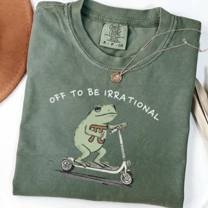 May include: Green Comfort Colors t-shirt featuring the text "OFF TO BE IRRATIONAL" and a cartoon frog on a scooter, with a Pi symbol. A silver necklace and a brown hat are also visible. The t-shirt is folded.