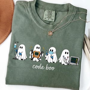 May include: An olive green crew neck t-shirt featuring a graphic of four white ghosts dressed as medical professionals with the text code boo. The first ghost holds an IV pole, the second a defibrillator, the third a syringe, and the fourth a monitor.
