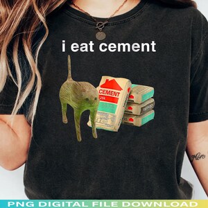 May include: Dark gray t-shirt with the text "i eat cement". The shirt features a cartoon cat and a bag of cement with the word "CEMENT" and a house graphic. There are also three smaller cement blocks.