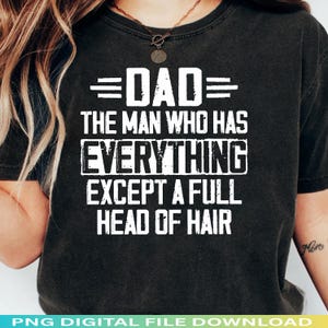 May include: Black t-shirt with white text that reads "DAD THE MAN WHO HAS EVERYTHING EXCEPT A FULL HEAD OF HAIR." The shirt has a crew neck and short sleeves. The text is centered on the front of the shirt.