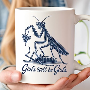 May include: White ceramic mug featuring a dark blue illustration of a praying mantis with the text "Girls will be Girls". The mug has a curved handle and is held by a person.