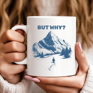May include: White ceramic mug with a blue mountain and penguin graphic. The mug has the text "BUT WHY?" in blue above the mountain illustration. The mug has a white handle and is held by a person.