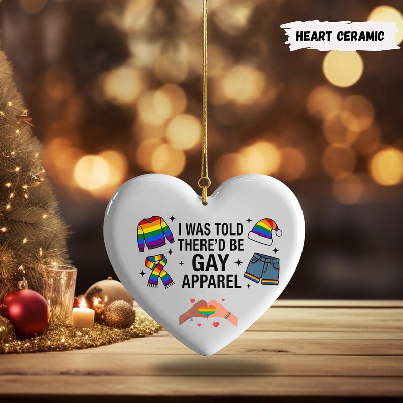 May include: A white heart-shaped ceramic ornament with a gold string for hanging. The ornament features the text "I WAS TOLD THERE'D BE GAY APPAREL" and illustrations of clothing items in rainbow colors. The words "HEART CERAMIC" are at the top.