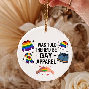 May include: A white ceramic ornament with a gold string, featuring the text "I WAS TOLD THERE'D BE GAY APPAREL." The design includes rainbow-colored clothing illustrations and a heart-shaped hand gesture.