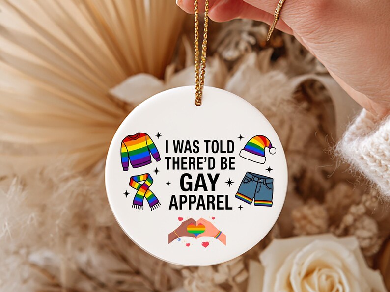May include: A white ceramic ornament with a gold string, featuring the text "I WAS TOLD THERE'D BE GAY APPAREL." The design includes rainbow-colored clothing illustrations and a heart-shaped hand gesture.