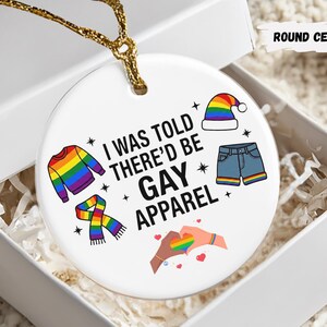 May include: A round ceramic ornament with the text "I WAS TOLD THERE'D BE GAY APPAREL" and colorful illustrations of clothing items. The ornament is in a white gift box with gold string.