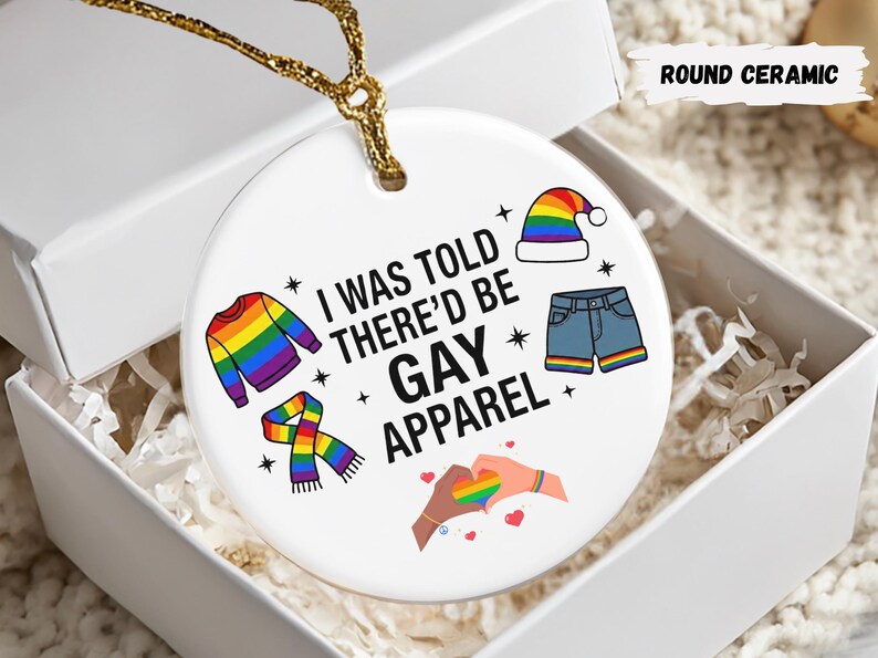 May include: A round ceramic ornament with the text "I WAS TOLD THERE'D BE GAY APPAREL" and colorful illustrations of clothing items. The ornament is in a white gift box with gold string.
