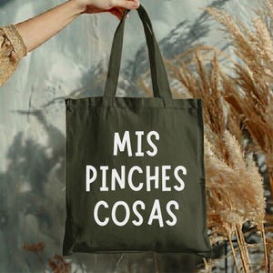 May include: An olive green tote bag with the words "MIS PINCHES COSAS" printed in white. The bag is held up by a hand, with a neutral background and dried plants on the right side.
