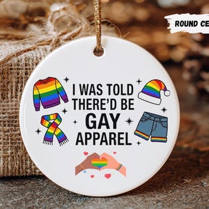 May include: A round ceramic ornament with a gold string for hanging. The ornament features the text "I WAS TOLD THERE'D BE GAY APPAREL" with colorful illustrations of clothing items and a rainbow heart design. The ornament is white.