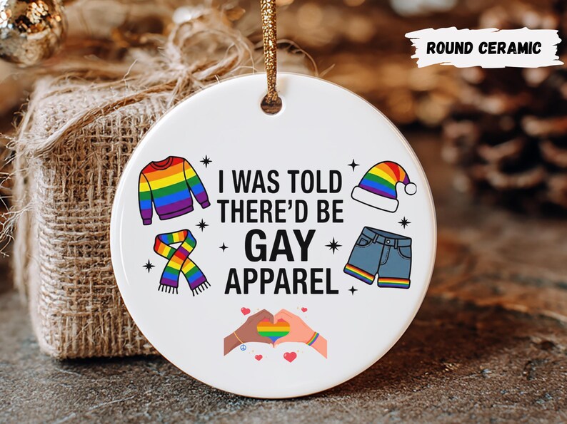 May include: A round ceramic ornament with a gold string for hanging. The ornament features the text "I WAS TOLD THERE'D BE GAY APPAREL" with colorful illustrations of clothing items and a rainbow heart design. The ornament is white.