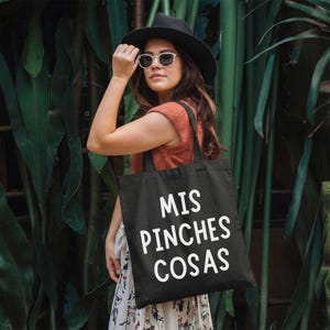 May include: Black tote bag with the text "MIS PINCHES COSAS" in white. The bag is carried by a person wearing a black hat, sunglasses, and an orange top. The background features lush green foliage.