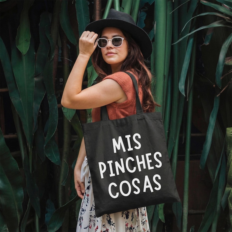 May include: Black tote bag with the text "MIS PINCHES COSAS" in white. The bag is carried by a person wearing a black hat, sunglasses, and an orange top. The background features lush green foliage.