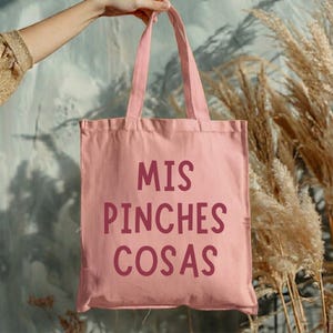 May include: A pink tote bag with the words "MIS PINCHES COSAS" printed in a darker pink. The bag is held up by a person. The background includes dried plants and a wall.