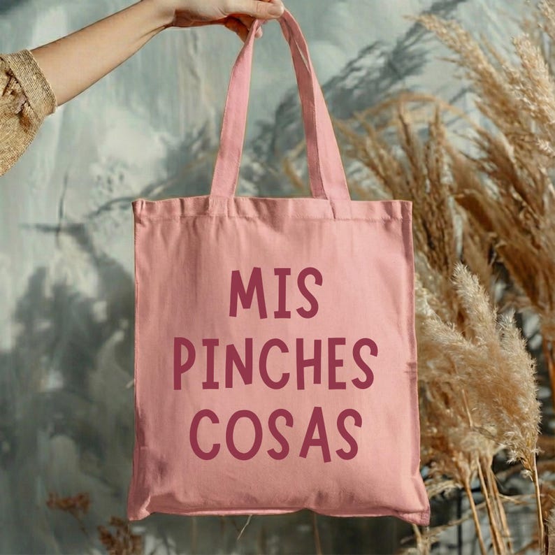 May include: A pink tote bag with the words "MIS PINCHES COSAS" printed in a darker pink. The bag is held up by a person. The background includes dried plants and a wall.