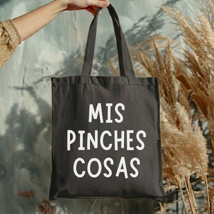 May include: A black tote bag with the words "MIS PINCHES COSAS" printed in white. The bag is held up by a person's hand. The background features dried plants and a textured wall.