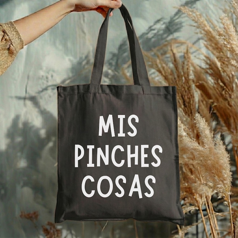May include: A black tote bag with the words "MIS PINCHES COSAS" printed in white. The bag is held up by a person's hand. The background features dried plants and a textured wall.