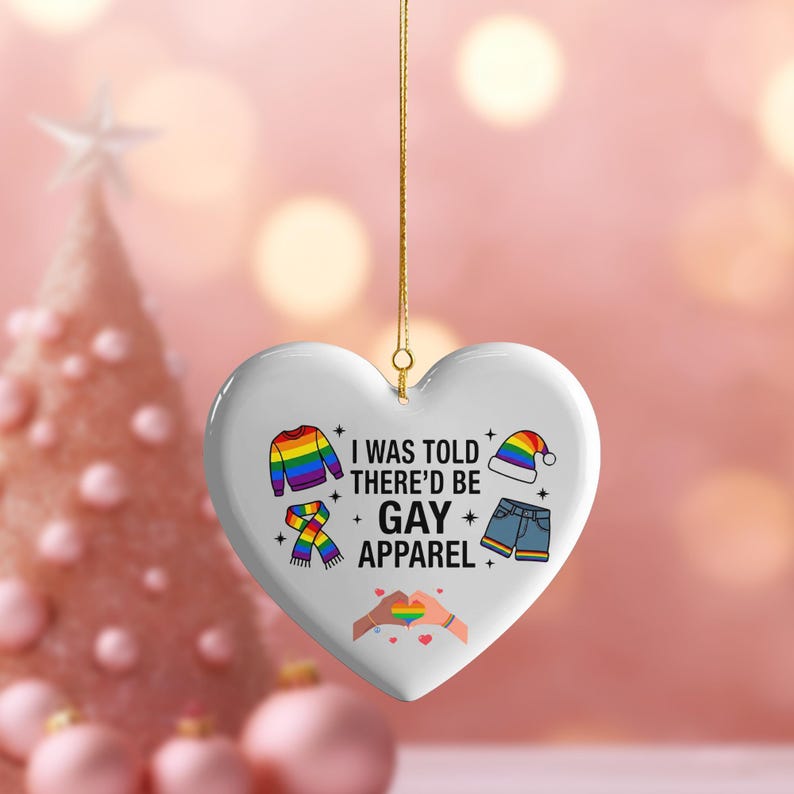 May include: White heart-shaped ornament with gold string, featuring a rainbow sweater, scarf, hat, and shorts. Text reads "I WAS TOLD THERE'D BE GAY APPAREL" with a rainbow heart symbol. Festive and colorful.