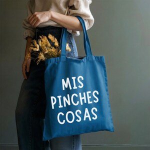 May include: A blue tote bag with white text that reads "MIS PINCHES COSAS." The bag is held by a person wearing jeans and a cream-colored sweater. The bag contains dried flowers.