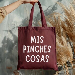 May include: A maroon tote bag with white text that reads "MIS PINCHES COSAS." The bag has two handles and is held up by a hand. The background features dried plants and a muted teal wall.