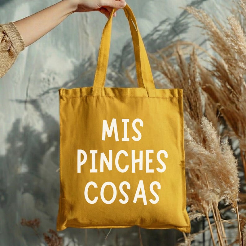 May include: Mustard yellow tote bag with the text "MIS PINCHES COSAS" in white. The bag is held up by a person's hand. The background features dried plants and a gray wall.