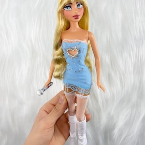 May include: A fashion doll with long blonde hair, wearing a light blue dress with a heart-shaped cutout and rhinestone embellishments. The doll is also wearing white boots and holding a microphone. The doll's dress is trimmed with lace.