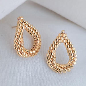 Puede incluir: Un par de pendientes dorados en forma de lágrima. Cada pendiente está construido con pequeñas cuentas rectangulares, creando un diseño texturizado de centro abierto. Los pendientes se muestran sobre una superficie blanca.