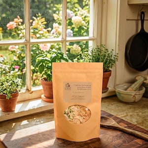 Puede incluir: Una bolsa de papel marrón de mezcla de sopa cremosa de patata y puerro está sobre una tabla de cortar de madera. La bolsa tiene una ventana transparente que muestra la mezcla de sopa. El fondo incluye una ventana con plantas y una cuchara de madera.