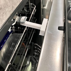 Może przedstawiać: Biała blokada drzwi zmywarki w kształcie litery T zamontowana na zmywarce ze stali nierdzewnej. Blokada ma zapobiegać otwieraniu drzwi. Widoczne jest wnętrze zmywarki z koszami i naczyniami.