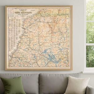 May include: A framed vintage map of the White Mountains region, featuring detailed cartography in shades of beige, blue, and brown. The map is displayed in a light-colored wooden frame, adding a classic touch to the wall decor. The map is hanging above a gray sofa.