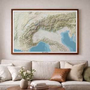 Könnte beinhalten: Eine gerahmte 3D-Reliefkarte der Alpen mit detaillierter Topografie in Beige-, Grün- und Blautönen. Die Karte ist vor einem weißen Hintergrund platziert und in dunklem Holz gerahmt. Der Text "DIE ALPEN" ist unten sichtbar.