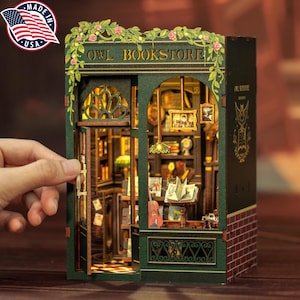 May include: A miniature Owl Bookstore model with a green facade, floral accents, and the text "OWL BOOKSTORE." The interior features bookshelves, framed pictures, and a warm, inviting glow. The model is held by a hand.