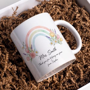 May include: White ceramic mug featuring a pastel rainbow and floral design. The mug has the text "Mrs. Smith, Thank you for helping me grow from Sophie" in black script. The mug is in a white gift box with brown paper filler.