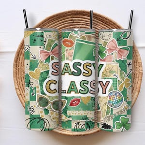 May include: Three stainless steel tumblers with black straws, decorated with green and white checkered patterns and various stickers. The tumblers have the words "SASSY CLASSY" in the center, with images of bows, sunglasses, and shamrocks.
