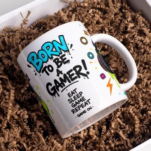 May include: White ceramic mug with the text "BORN TO BE A GAMER!" in blue and black graffiti-style lettering. The mug also features the phrases "EAT SLEEP GAME REPEAT" and "GAME ON." Colourful accents and a lightning bolt design complete the look.
