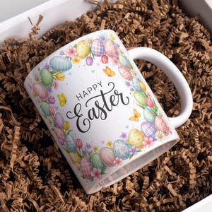 Op de afbeelding: Witte keramische mok met een ontwerp van pastelkleurige paaseieren, bloemen en vlinders. De woorden "Happy Easter" zijn in zwart schrift geschreven. De mok ligt in bruin verpakkingsmateriaal.
