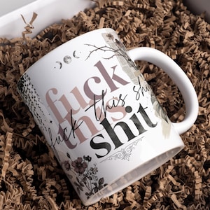 May include: White ceramic mug with the phrase "fuck this shit" in a mix of pink and black fonts. The mug features floral and celestial accents, and is set in a box with brown paper filler.