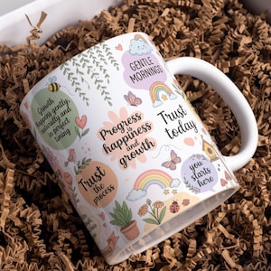 May include: White ceramic mug with a wraparound design featuring pastel illustrations of a bee, butterflies, rainbows, and flowers. Text includes phrases like "Gentle Mornings" and "Trust today."