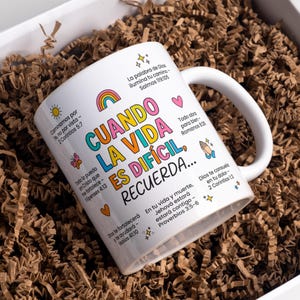 Może przedstawiać: Biały ceramiczny kubek z zakrzywionym uchwytem, z kolorowym tekstem i ilustracjami. Kubek ma tęczę, serca i gwiazdy, ze słowami "CUANDO LA VIDA ES DIFICIL, RECUERDA..." i wersetami biblijnymi. Kubek jest prezentowany w pudełku z brązowym papierem.