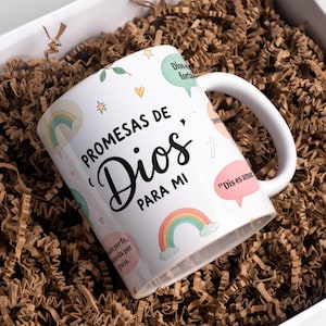 May include: White ceramic mug with the text "PROMESAS DE Dios PARA MI" and various colorful illustrations, including rainbows and speech bubbles. The mug is inside a white box filled with brown crinkle paper.